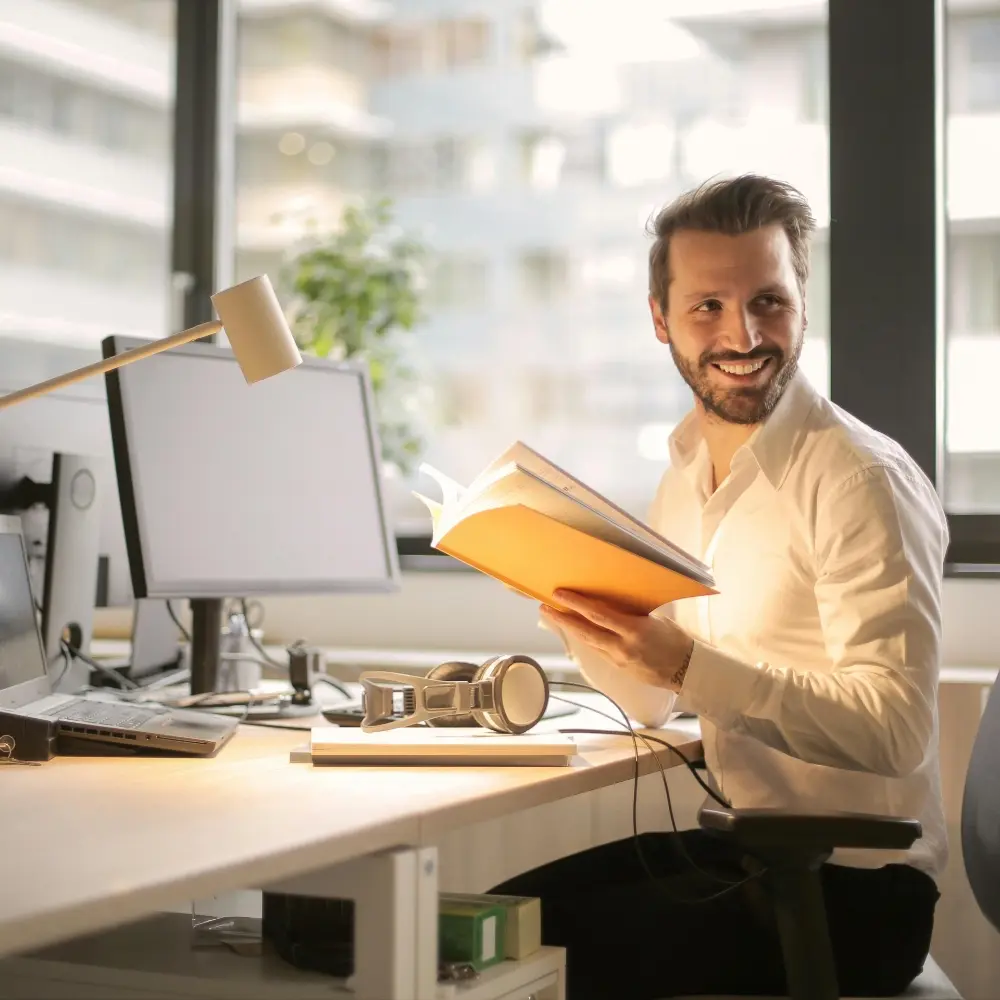 A smiling man with a beard sits at a modern office desk, looking away from his workstation and holding an open book with an orange cover. He is wearing a white button-down shirt. His desk is equipped with a laptop, a large monitor, a desk lamp, and a pair of headphones. Large windows in the background reveal an urban office setting with soft, natural light.